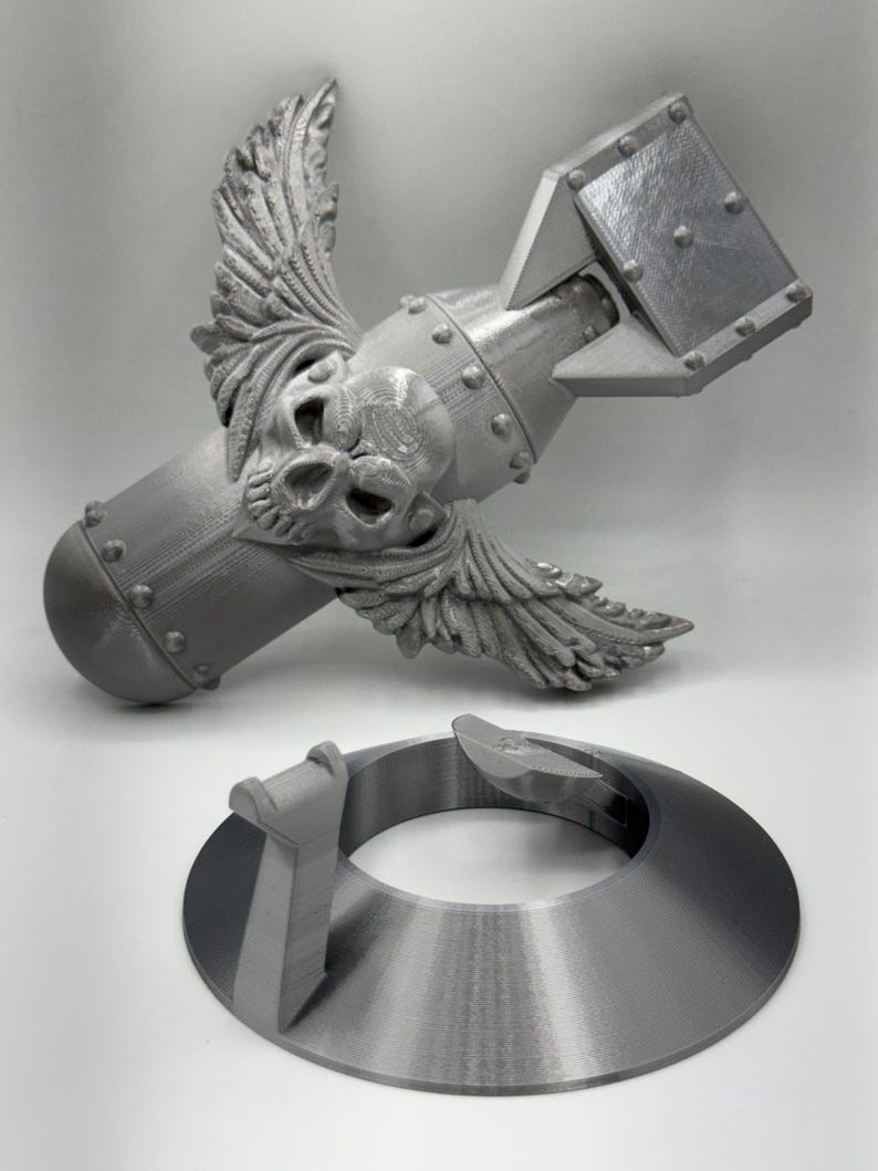 May include: A gray, 3D-printed model of a bomb with a skull and wings, and a separate circular base. The bomb has a detailed skull design and wing accents. The base is a ring with a raised section.