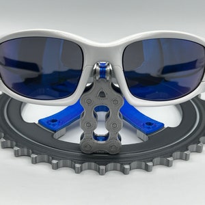 Pu&ograve; includere: Occhiali da sole bianchi con lenti blu sono esposti su un supporto per piatto catena da bicicletta grigio. Il supporto &egrave; in plastica stampata in 3D e presenta una catena blu.