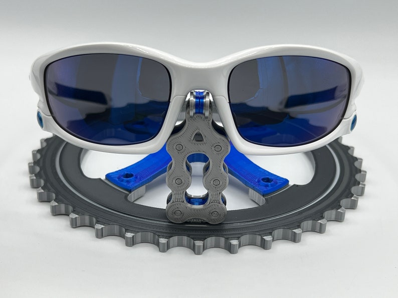 Pu&ograve; includere: Occhiali da sole bianchi con lenti blu sono esposti su un supporto per piatto catena da bicicletta grigio. Il supporto &egrave; in plastica stampata in 3D e presenta una catena blu.