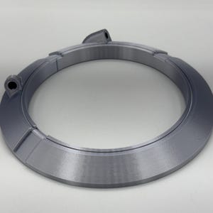 May include: A gray, circular object with a segmented design. The object has a raised, angled outer edge and two small, cylindrical protrusions. The overall shape is a ring, possibly a component or decorative element.