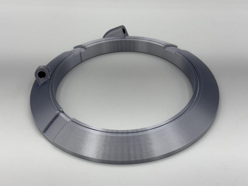 May include: A gray, circular object with a segmented design. The object has a raised, angled outer edge and two small, cylindrical protrusions. The overall shape is a ring, possibly a component or decorative element.