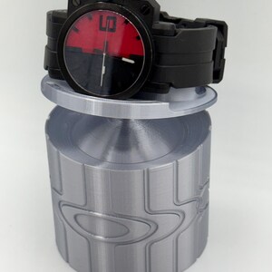 May include: Black wristwatch with a red and black face, displaying the number 6. The watch is on a silver cylindrical stand with a repeating design. The watch band is black.