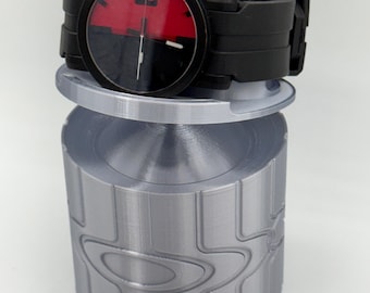 May include: Black wristwatch with a red and black face, displaying the number 6. The watch is on a silver cylindrical stand with a repeating design. The watch band is black.