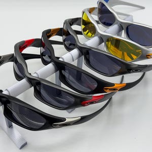May include: An assortment of sunglasses with dark lenses and various frame colors, including black, silver, and yellow. The sunglasses are displayed on a white stand, showcasing their design and style. The frames feature accents of red, orange, and blue.
