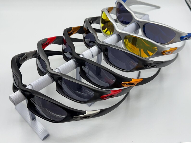 May include: An assortment of sunglasses with dark lenses and various frame colors, including black, silver, and yellow. The sunglasses are displayed on a white stand, showcasing their design and style. The frames feature accents of red, orange, and blue.
