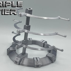 May include: A silver-colored, three-tiered display stand with a circular base. The stand has three curved arms on each tier, designed to hold items like swords or other collectibles. The stand is labeled "TRIPLE TIER" at the top.