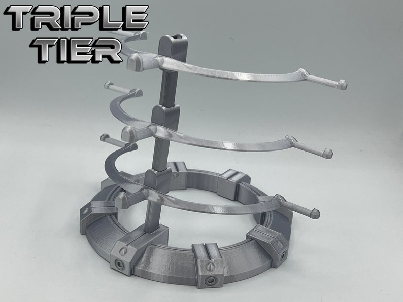 May include: A silver-colored, three-tiered display stand with a circular base. The stand has three curved arms on each tier, designed to hold items like swords or other collectibles. The stand is labeled "TRIPLE TIER" at the top.