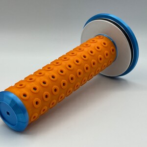 May include: Orange and blue motorcycle handlebar grip with a textured surface and a circular pattern of holes. The grip has a blue end cap and a white end cap with a black ring.
