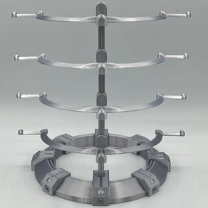 May include: A silver metal stand with three tiers for displaying items. Each tier has a curved, open design with two arms on each side. The stand has a circular base with a textured surface.