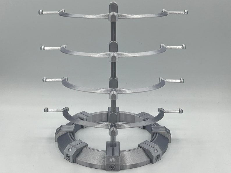 May include: A silver metal stand with three tiers for displaying items. Each tier has a curved, open design with two arms on each side. The stand has a circular base with a textured surface.