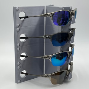 May include: A gray metal stand with four slots for holding sunglasses. The stand has a textured surface and is designed to hold sunglasses by the arms. Four pairs of sunglasses are displayed on the stand, with lenses in shades of blue and brown.