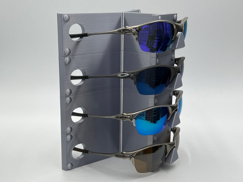 May include: A gray metal stand with four slots for holding sunglasses. The stand has a textured surface and is designed to hold sunglasses by the arms. Four pairs of sunglasses are displayed on the stand, with lenses in shades of blue and brown.