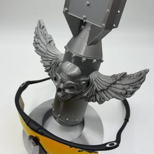May include: Black sunglasses with yellow lenses rest on a gray, stylized bomb-shaped sculpture. The sculpture features a skull with wings. The sunglasses have a black frame and a brown nose bridge. The sculpture is on a circular base.