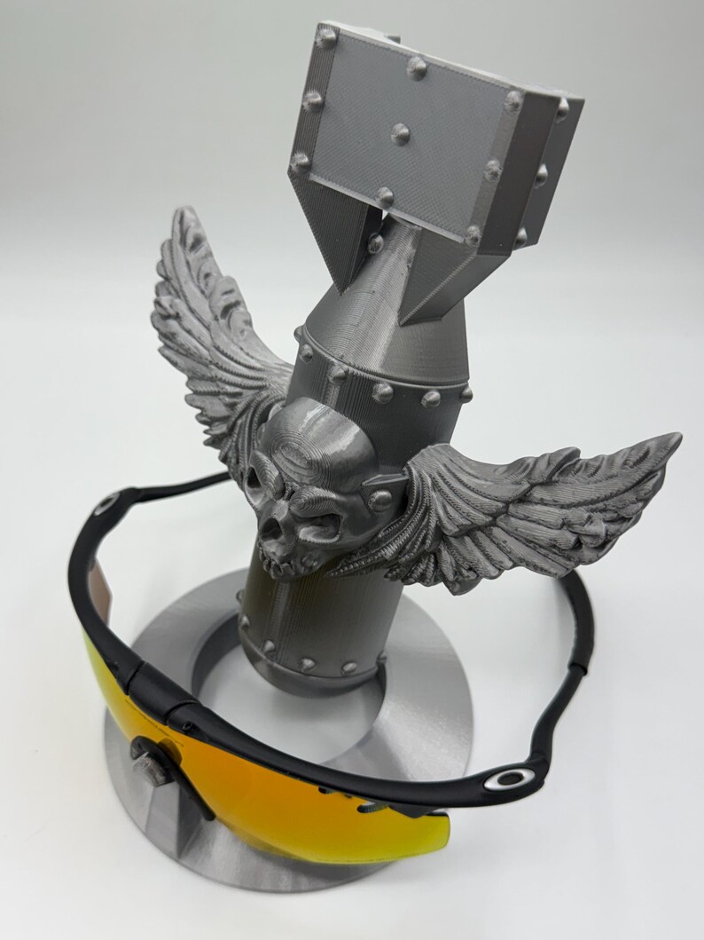 May include: Black sunglasses with yellow lenses rest on a gray, stylized bomb-shaped sculpture. The sculpture features a skull with wings. The sunglasses have a black frame and a brown nose bridge. The sculpture is on a circular base.