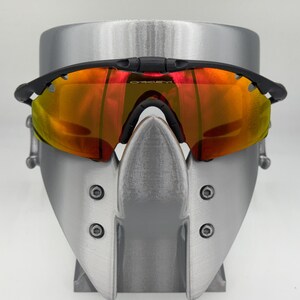 May include: Black sports sunglasses with orange and yellow lenses. The sunglasses are displayed on a silver stand. The lenses have a reflective quality, and the frame is black.