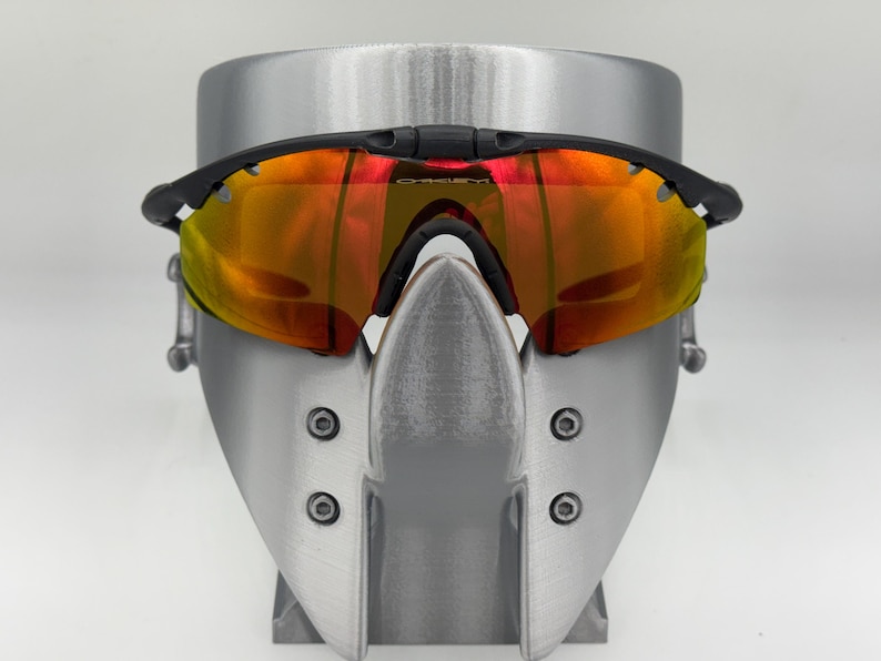 May include: Black sports sunglasses with orange and yellow lenses. The sunglasses are displayed on a silver stand. The lenses have a reflective quality, and the frame is black.