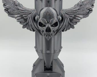 May include: A gray, 3D-printed sculpture of a bomb with a skull and wings design. The bomb has a cylindrical body with rivets and a pointed base. The skull is centered on the body, with detailed wings extending outward.