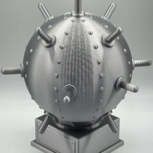 May include: A metallic grey, spherical object with protruding spikes and studs, mounted on a square base. The object has a textured surface and a detailed, industrial design, resembling a historical naval mine.