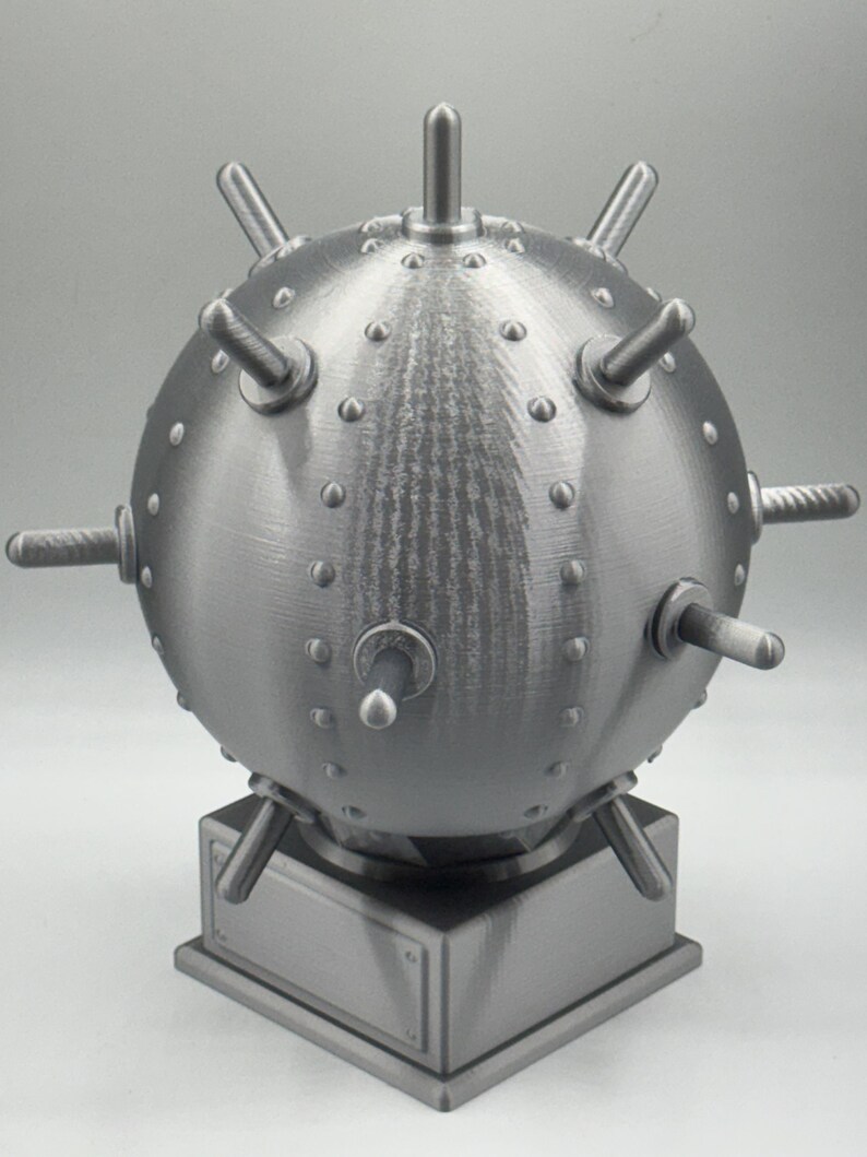May include: A metallic grey, spherical object with protruding spikes and studs, mounted on a square base. The object has a textured surface and a detailed, industrial design, resembling a historical naval mine.