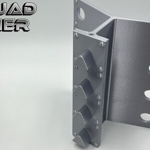 May include: A gray metal object with four triangular protrusions on the left side and three rows of holes on the right side. The object is labeled "QUAD TIER".