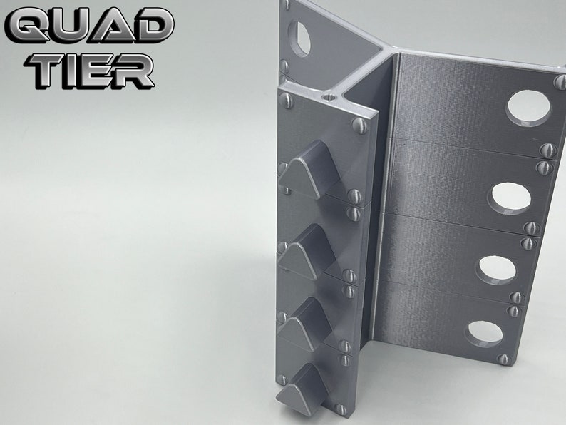May include: A gray metal object with four triangular protrusions on the left side and three rows of holes on the right side. The object is labeled "QUAD TIER".