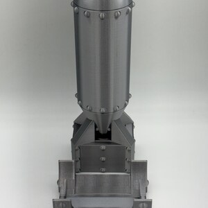May include: A detailed, gray, 3D-printed model of a bomb. The model features a rounded top, a cylindrical body, and a rectangular base. The surface has a textured, metallic appearance, with visible rivets and seams. The design is a replica of a historical bomb.