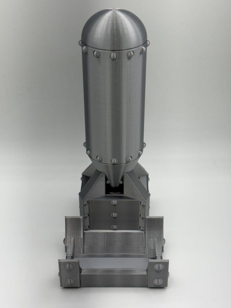 May include: A detailed, gray, 3D-printed model of a bomb. The model features a rounded top, a cylindrical body, and a rectangular base. The surface has a textured, metallic appearance, with visible rivets and seams. The design is a replica of a historical bomb.