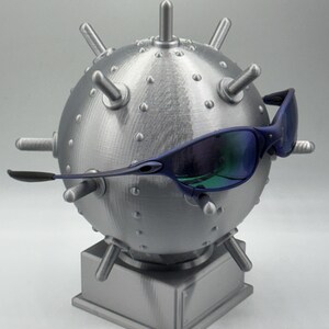 May include: A grey, spherical sculpture, reminiscent of a mine, featuring protruding spikes and rivets. The sculpture is mounted on a square base and is accessorized with blue-tinted sunglasses. The sunglasses have a sleek, modern design.