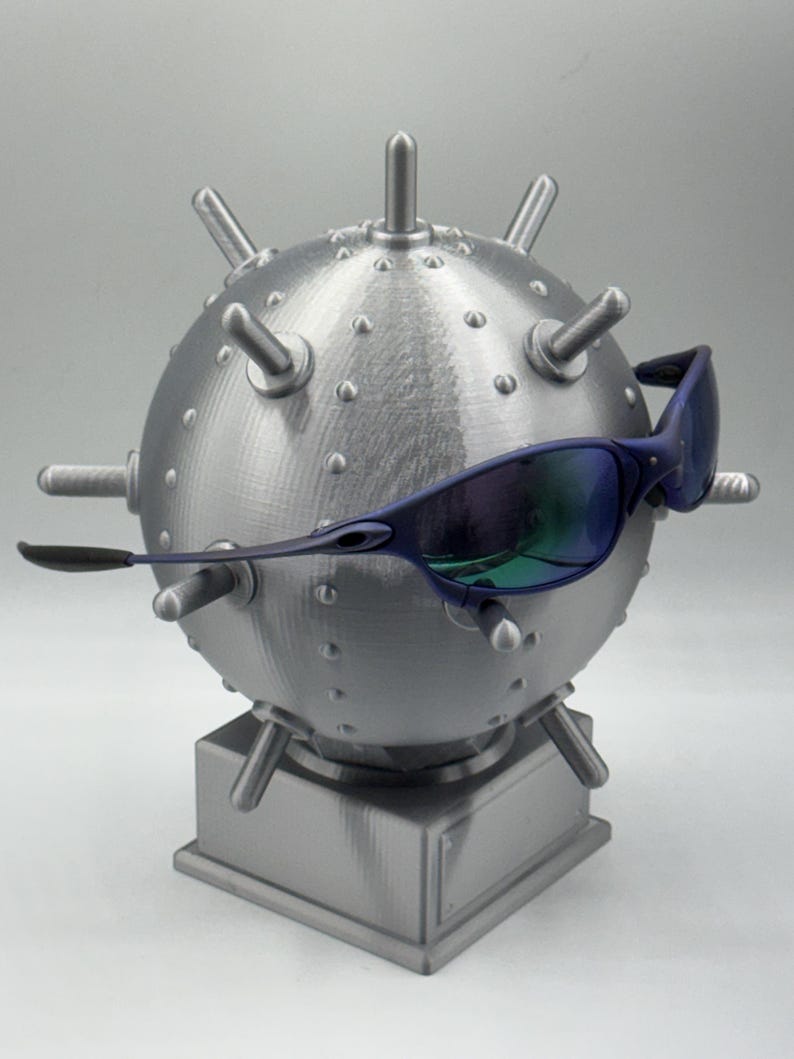 May include: A grey, spherical sculpture, reminiscent of a mine, featuring protruding spikes and rivets. The sculpture is mounted on a square base and is accessorized with blue-tinted sunglasses. The sunglasses have a sleek, modern design.