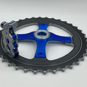 Pu&ograve; includere: Un ingranaggio stampato in 3D con finitura blu e argento. L'ingranaggio ha un design a quattro razze e una catena attaccata al lato.