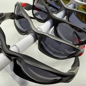 May include: A collection of sunglasses with dark lenses and black frames. Several pairs are visible, with a focus on the sleek, curved design of the eyewear. Some frames have red or silver accents. The sunglasses are displayed on a white surface.