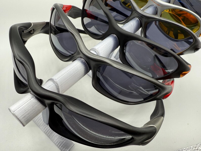 May include: A collection of sunglasses with dark lenses and black frames. Several pairs are visible, with a focus on the sleek, curved design of the eyewear. Some frames have red or silver accents. The sunglasses are displayed on a white surface.