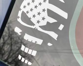 May include: A white skull sticker with an American flag design on a car window. The skull is outlined in black and has a white star pattern.