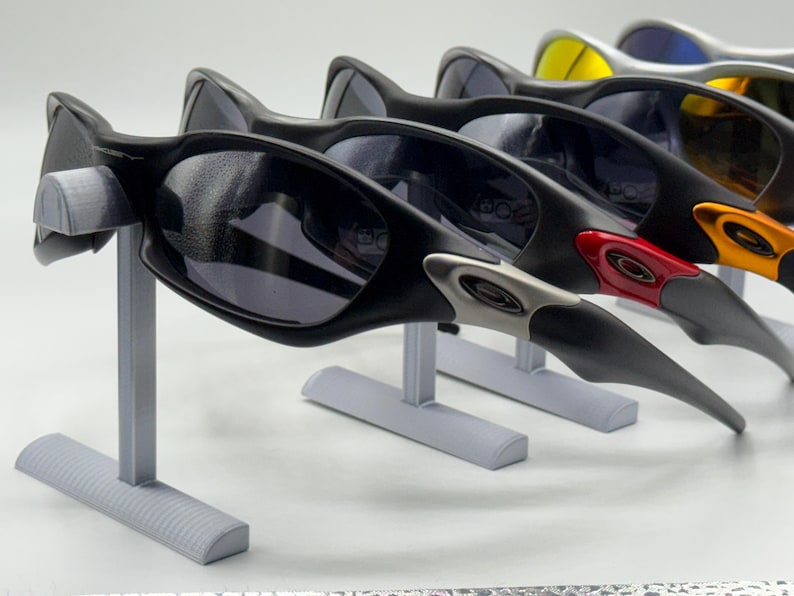 May include: A display of several pairs of sunglasses with black frames and dark lenses. The sunglasses are on gray stands. Some of the sunglasses have colored accents, including red, orange, and silver.