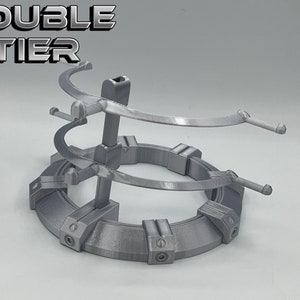 May include: A silver metal double-tier stand with a circular base and two curved arms on each tier. The stand is designed to hold items like figurines or other collectibles. The text "DOUBLE TIER" is printed on the top of the stand.