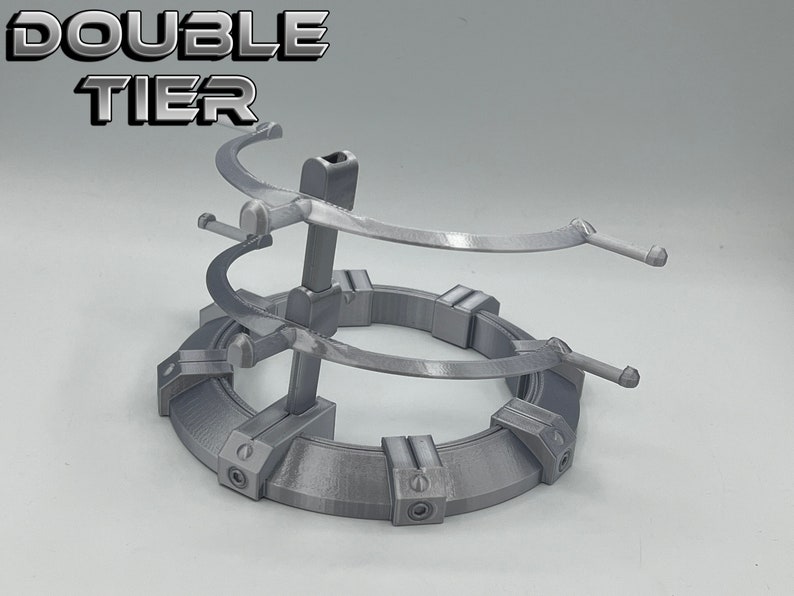 May include: A silver metal double-tier stand with a circular base and two curved arms on each tier. The stand is designed to hold items like figurines or other collectibles. The text "DOUBLE TIER" is printed on the top of the stand.