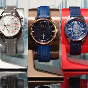 May include: Three wristwatches on display stands. The watch on the left has a white face with black numbers and a gray leather band. The middle watch has a blue face with black hands and a blue leather band. The watch on the right has a blue face with white numbers and a blue fabric band.