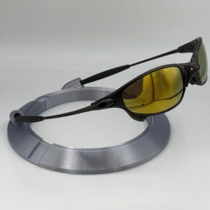 May include: Black sunglasses with gold-tinted lenses rest on a gray, circular display stand. The sunglasses have a sleek, modern design with a curved frame and arms. The stand is a light gray color.