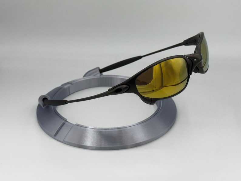 May include: Black sunglasses with gold-tinted lenses rest on a gray, circular display stand. The sunglasses have a sleek, modern design with a curved frame and arms. The stand is a light gray color.