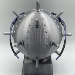 May include: A metallic grey, spherical ornament with numerous protruding spikes and a rectangular base. The sphere is covered in small, evenly spaced rivets. Blue and black curved elements are attached to the sides. A decorative item, possibly a sculpture or display piece.