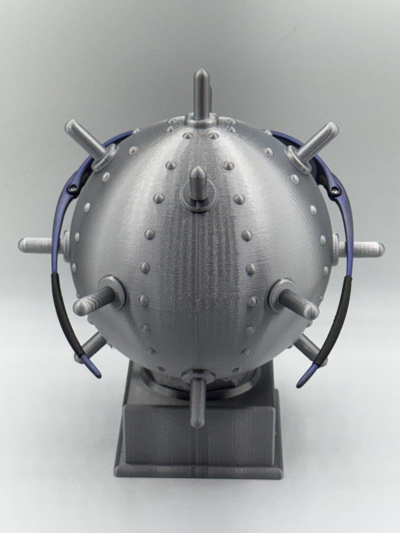 May include: A metallic grey, spherical ornament with numerous protruding spikes and a rectangular base. The sphere is covered in small, evenly spaced rivets. Blue and black curved elements are attached to the sides. A decorative item, possibly a sculpture or display piece.
