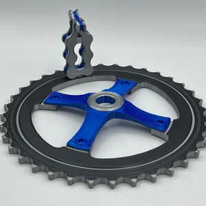 Pu&ograve; includere: Un pignone per bicicletta stampato in 3D con finitura blu e argento. Il pignone ha un design a forma di stella con quattro bracci e un foro centrale. Un piccolo guida catena stampato in 3D blu &egrave; attaccato alla parte superiore del pignone.