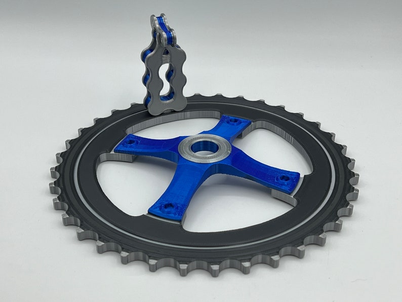 Pu&ograve; includere: Un pignone per bicicletta stampato in 3D con finitura blu e argento. Il pignone ha un design a forma di stella con quattro bracci e un foro centrale. Un piccolo guida catena stampato in 3D blu &egrave; attaccato alla parte superiore del pignone.