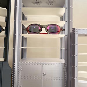 K&ouml;nnte beinhalten: Ein silberner Schaukasten mit mehreren Regalen, zur Aufnahme von Brillen. Der Schrank hat ein genietetes Metallaussehen und einen kleinen Schrank am Boden. Eine Sonnenbrille mit schwarzem Rahmen und roten Akzenten liegt auf einem Regal.