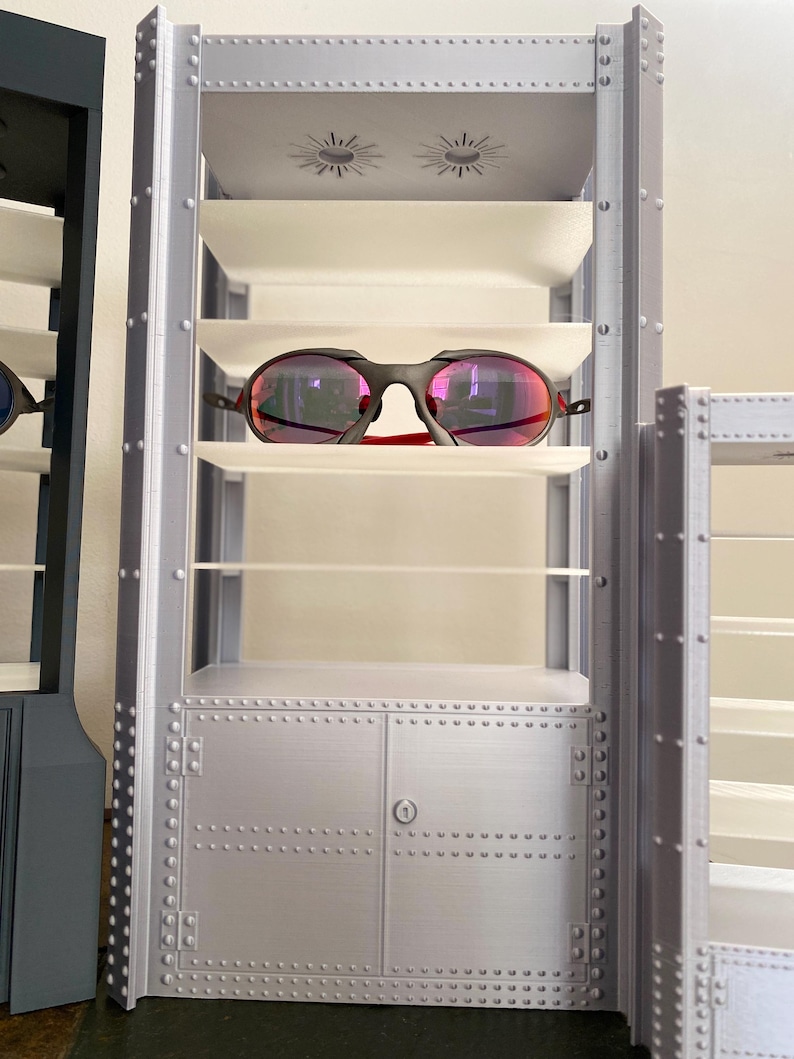 K&ouml;nnte beinhalten: Ein silberner Schaukasten mit mehreren Regalen, zur Aufnahme von Brillen. Der Schrank hat ein genietetes Metallaussehen und einen kleinen Schrank am Boden. Eine Sonnenbrille mit schwarzem Rahmen und roten Akzenten liegt auf einem Regal.