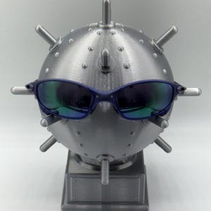 May include: A silver-coloured, spherical sculpture resembling a mine, with protruding spikes and a rectangular base. The sculpture has blue-framed sunglasses resting on it, with reflective lenses that show green and blue hues.