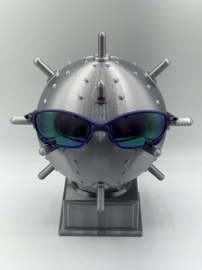 May include: A silver-coloured, spherical sculpture resembling a mine, with protruding spikes and a rectangular base. The sculpture has blue-framed sunglasses resting on it, with reflective lenses that show green and blue hues.