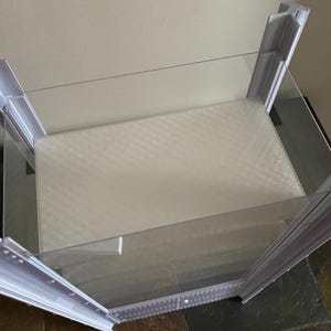 K&ouml;nnte beinhalten: Eine transparente, rechteckige Vitrine mit wei&szlig;em Innenraum und einer schr&auml;gen, transparenten Oberseite. Der Rahmen ist wei&szlig; und hat eine Reihe kleiner L&ouml;cher an den Kanten. Die Vitrine dient zur Pr&auml;sentation von Artikeln.