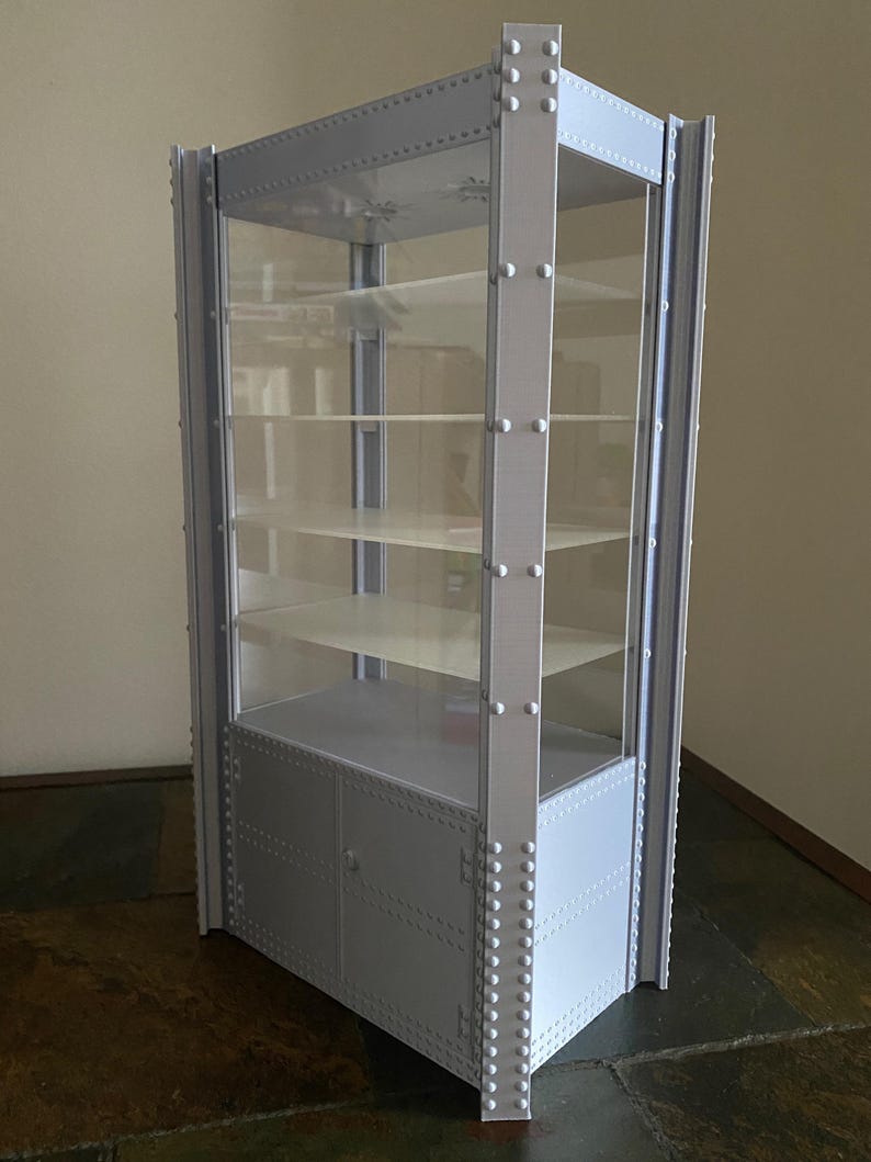K&ouml;nnte beinhalten: Ein grauer Vitrinenschrank mit Glasb&ouml;den und T&uuml;ren. Der Schrank hat einen genieteten Metallrahmen und eine rechteckige Form. Er ist f&uuml;r die Pr&auml;sentation von Sammlerst&uuml;cken oder anderen Gegenst&auml;nden konzipiert.