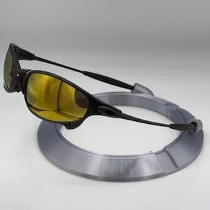 May include: Black sunglasses with gold-tinted lenses rest on a gray circular display stand. The sunglasses have a sleek, modern design with a black frame and arms. The lenses reflect a golden hue.